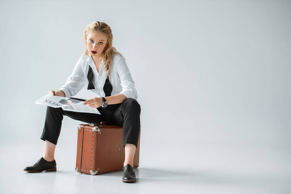 mooi stijlvol meisje business krant lezen zittend op vintage koffer op grijs - Foto, afbeelding