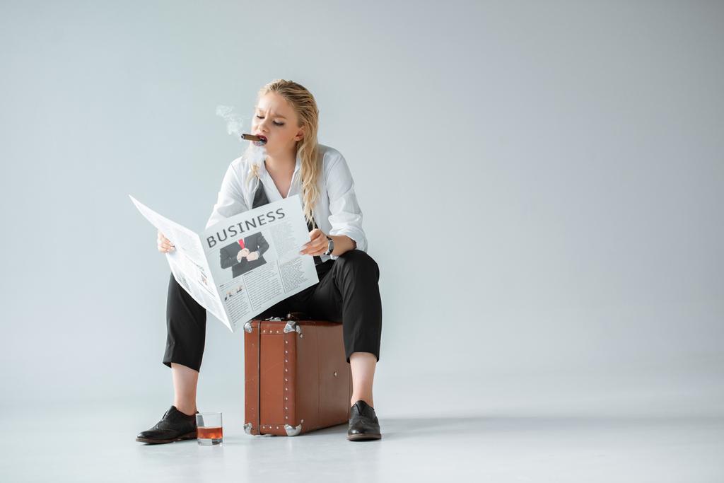 fille à la mode avec cigare et verre de whisky lecture journal d'affaires tout en étant assis sur une valise rétro sur gris
 - Photo, image
