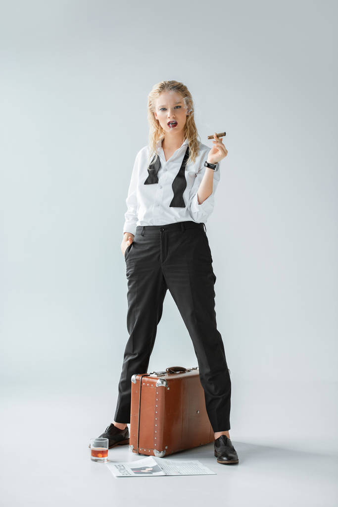 приваблива модна дівчина палить сигару, стоячи на сірому з старовинною сумкою для подорожей, газетою та келихом віскі
 - Фото, зображення