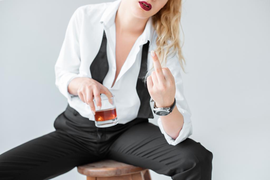 vue recadrée d'une femme élégante avec un verre de whisky assis sur un tabouret et montrant son majeur, isolé sur du gris
 - Photo, image