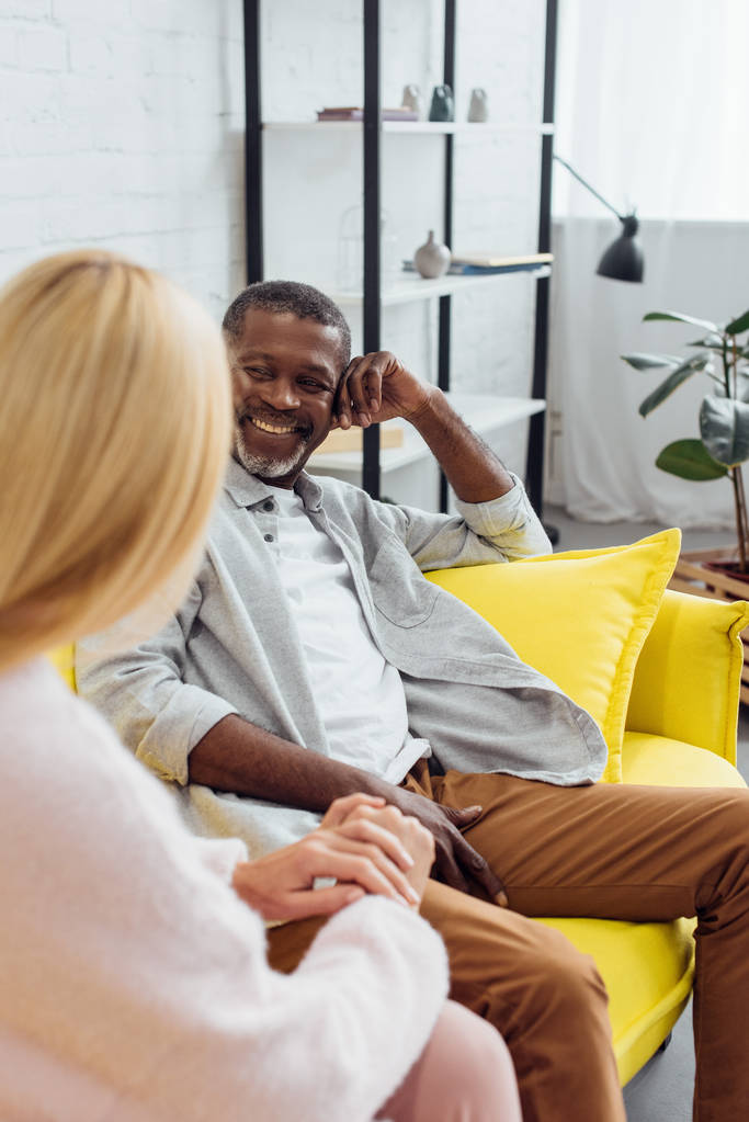 sorridente maduro afro-americano homem sentado no sofá e falando com mulher loira
  - Foto, Imagem