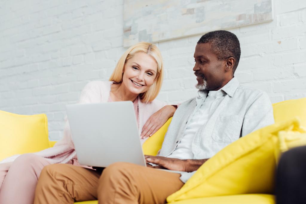 щаслива жінка сидить на дивані з афроамериканським чоловіком і використовує ноутбук
 - Фото, зображення