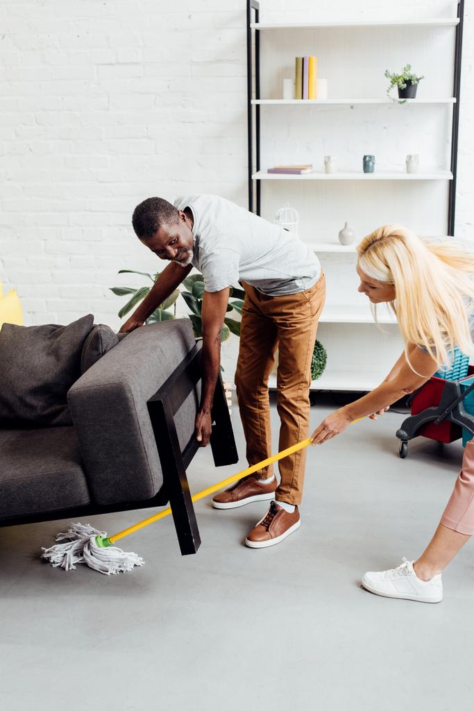 Blondine putzt Boden mit Wischmopp, während Afroamerikaner schweres Sofa hochhebt - Foto, Bild