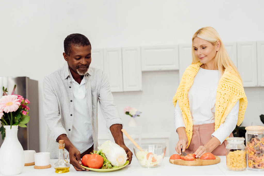 femme souriante tranchant des tomates sur la planche à découper tandis que l'homme afro-américain tenant assiette avec des légumes
 - Photo, image