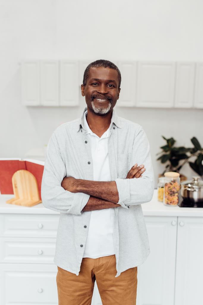 szczęśliwy african american dojrzały mężczyzna z ramionami skrzyżowane stojący w kuchni - Zdjęcie, obraz