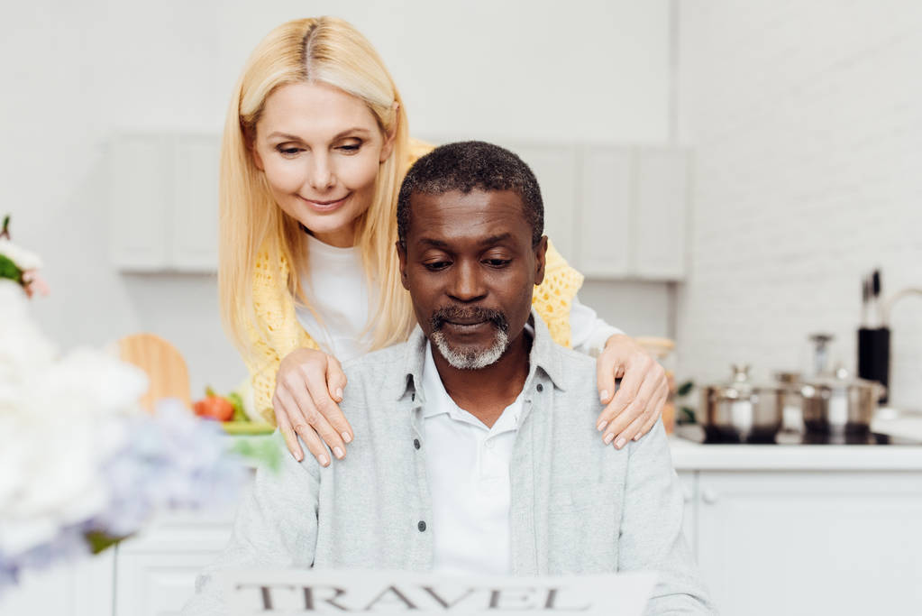 Ältere lächelnde Frau umarmt einen afrikanisch-amerikanischen Mann, während sie in der Küche die Reisezeitung liest - Foto, Bild