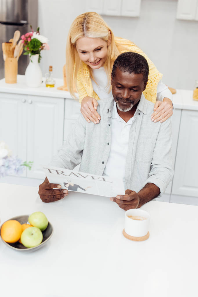 mature smiling woman embracing man while reading travel newspaper at kitchen - Photo, Image