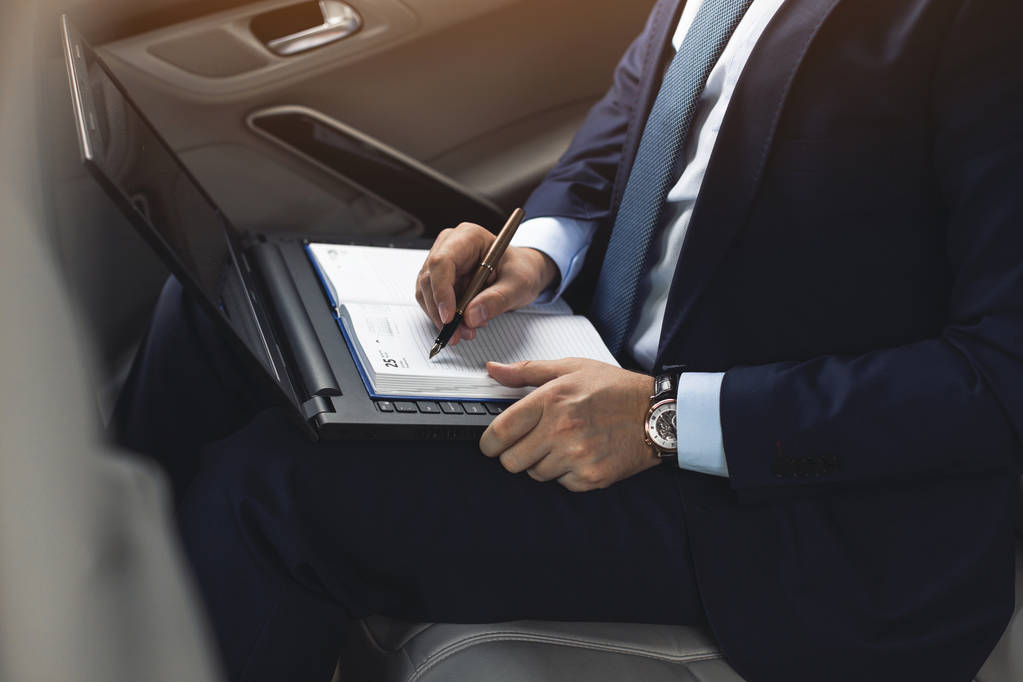 un joven hombre de negocios en un traje está sentado en el asiento trasero de un coche de negocios con un ordenador portátil y un cuaderno, negociaciones comerciales
 - Foto, imagen