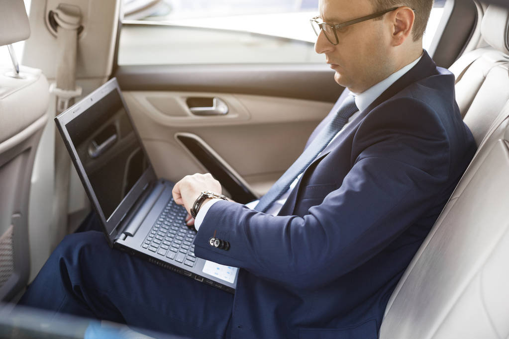 um jovem homem de negócios em um terno está sentado no banco de trás de um carro de negócios com um laptop e um notebook, negociações de negócios
 - Foto, Imagem