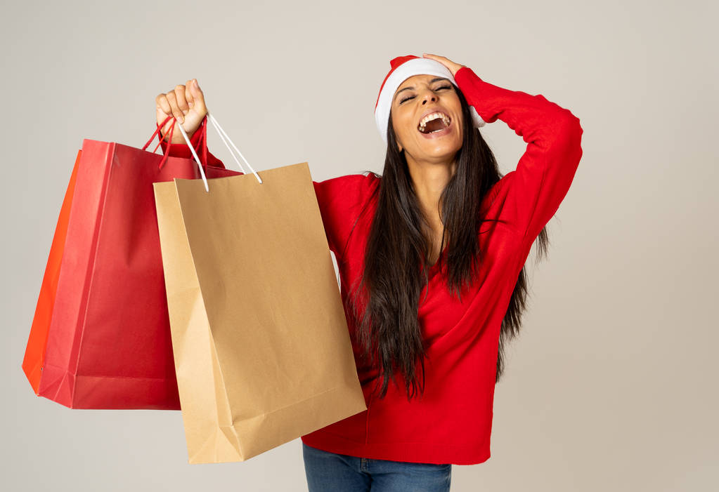 Nuori nainen valmis jouluun paperilla ostoskassit punainen ja Santa Claus hattu eristetty harmaa tausta on valmis joululahjat juhla kulutustavara ja myynti konsepti
. - Valokuva, kuva