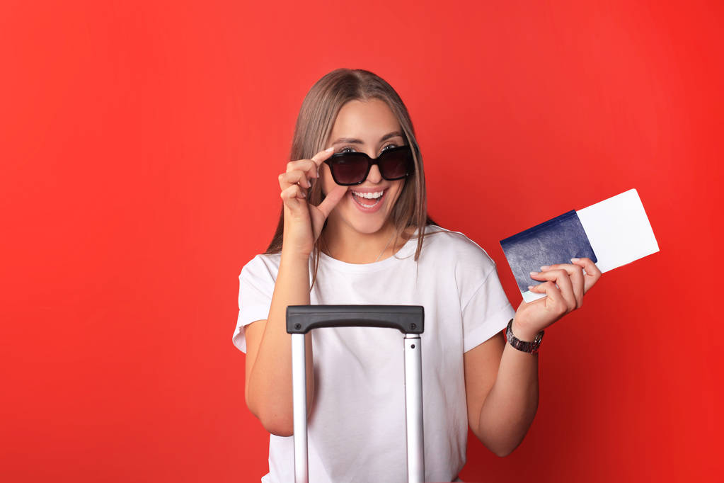 Молодая девушка турист в летней повседневной одежде, в солнечных очках, красный чемодан, паспорт изолирован на красном фоне
. - Фото, изображение
