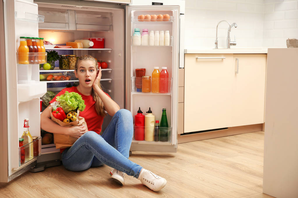 Emocjonalne młoda kobieta siedzi w pobliżu lodówki w kuchni - Zdjęcie, obraz