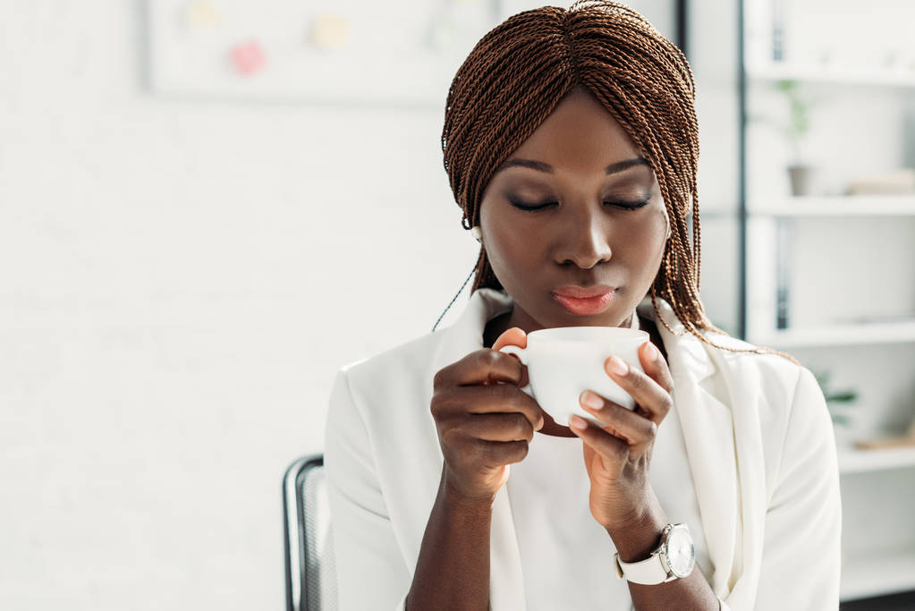 hermosa mujer de negocios adulta afroamericana en traje blanco con los ojos cerrados sentado en el escritorio, sosteniendo taza de café y aroma inhalatorio
 - Foto, Imagen