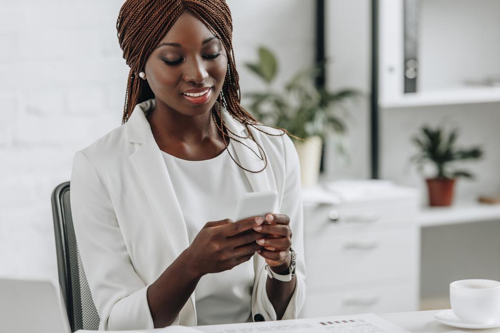 uśmiechający się african american kobieta dorosłych w białe stroje wizytowe, siedząc przy biurku i przy użyciu smartfona w biurze - Zdjęcie, obraz