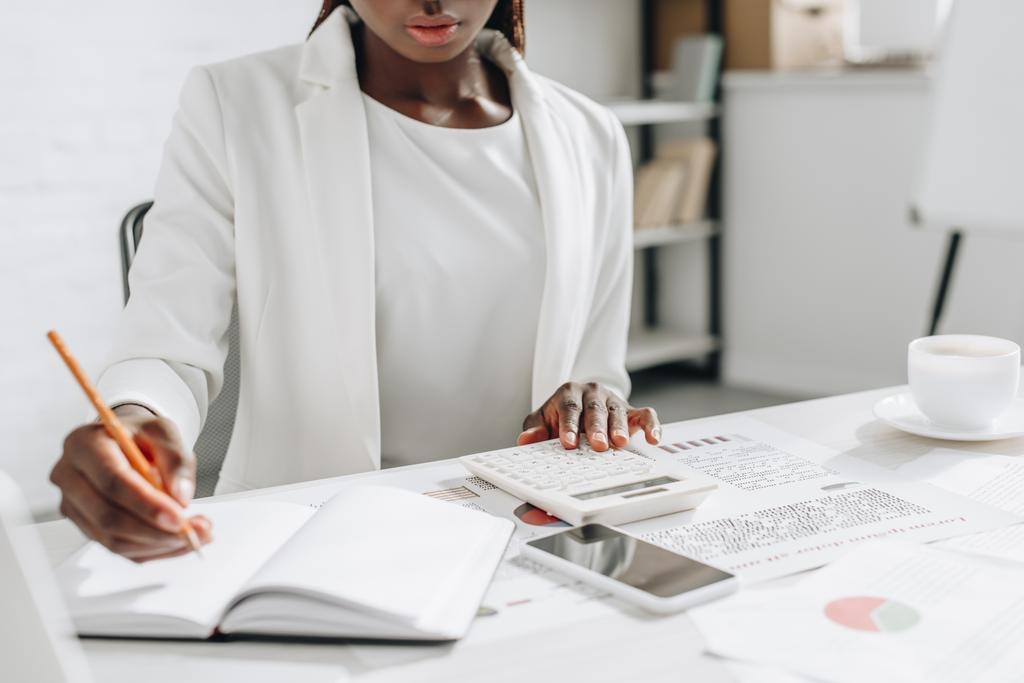 Nézd az afro-amerikai felnőtt üzletasszony övben fehér ruházat írás jegyzetfüzet és dolgozó irodai íróasztal - Fotó, kép