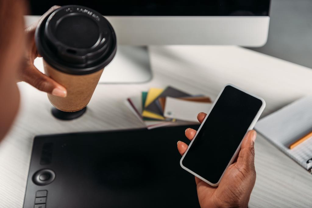 vista ritagliata di donna d'affari afro-americana in possesso di caffè per andare e smartphone con schermo bianco
 - Foto, immagini