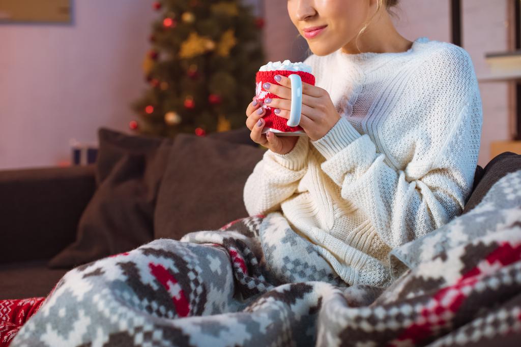 abgeschnittene Ansicht einer jungen Frau, die in eine Decke gehüllt auf einer Couch sitzt und eine Tasse heißen Kakao mit Marshmallows in der Weihnachtszeit hält  - Foto, Bild