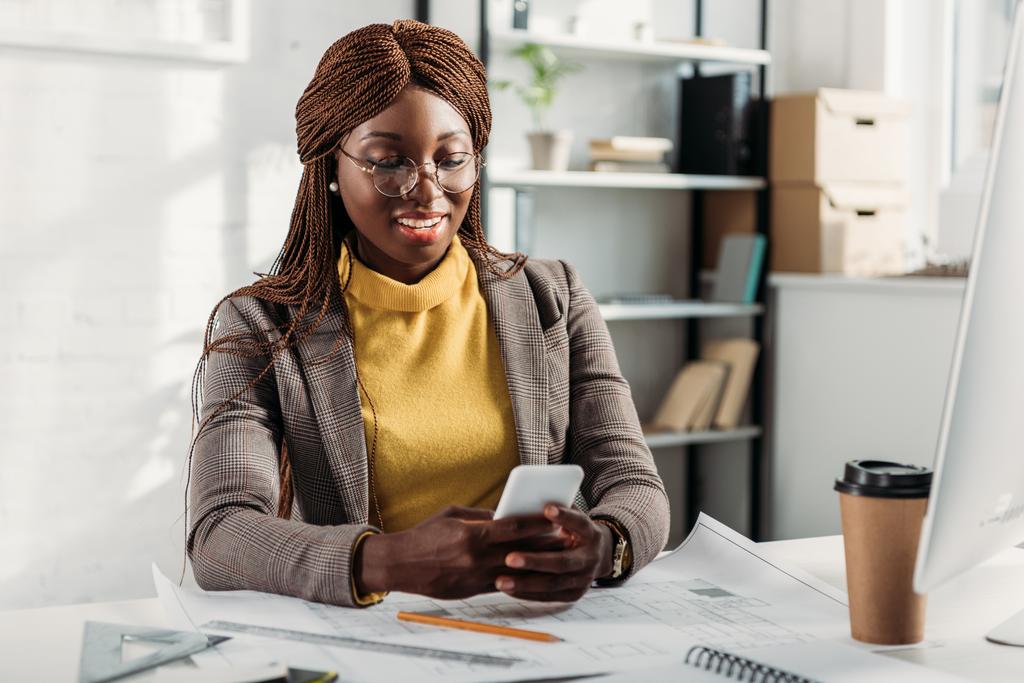 s úsměvem afroamerické ženy architekt v brýlích pomocí smartphone a práci u stolu s plány v úřadu - Fotografie, Obrázek