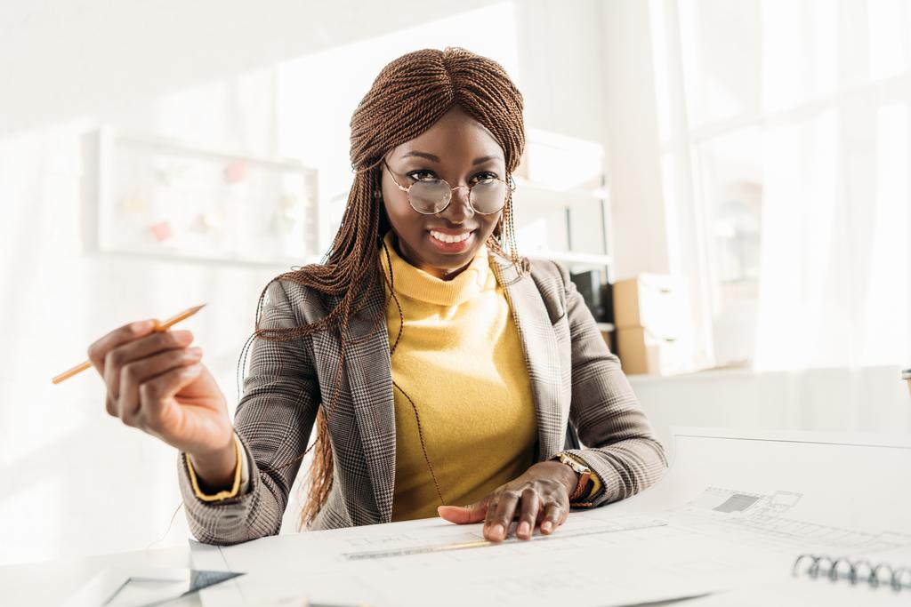 sorridente architetto afroamericana donna in bicchieri che tiene la matita, guarda la macchina fotografica e lavora al progetto alla scrivania con cianografie in ufficio
 - Foto, immagini
