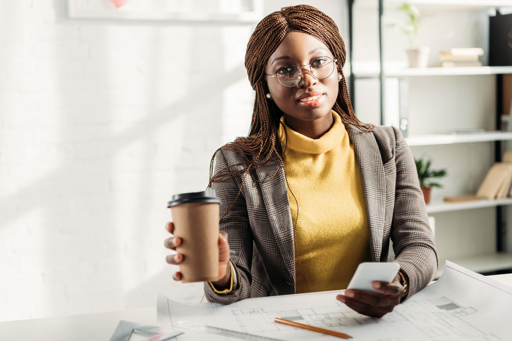 Αφρικανική αμερικανική γυναίκα αρχιτέκτονα σε ποτήρια κρατώντας καφέ να πάει, χρησιμοποιώντας το smartphone και εργάζονται στο γραφείο για το έργο με σχεδιαγράμματα - Φωτογραφία, εικόνα