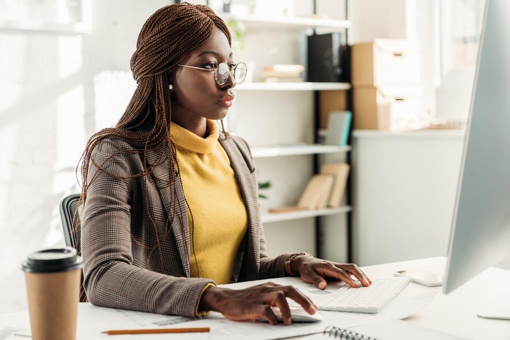 mujer de negocios afroamericana enfocada sentada en escritorio de computadora y trabajando en la oficina
 - Foto, imagen