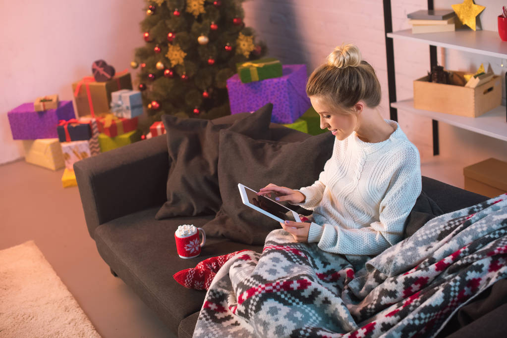 kanepede oturan ve tablet Noel zamanı kullanma gülümseyen genç sarışın kadın  - Fotoğraf, Görsel