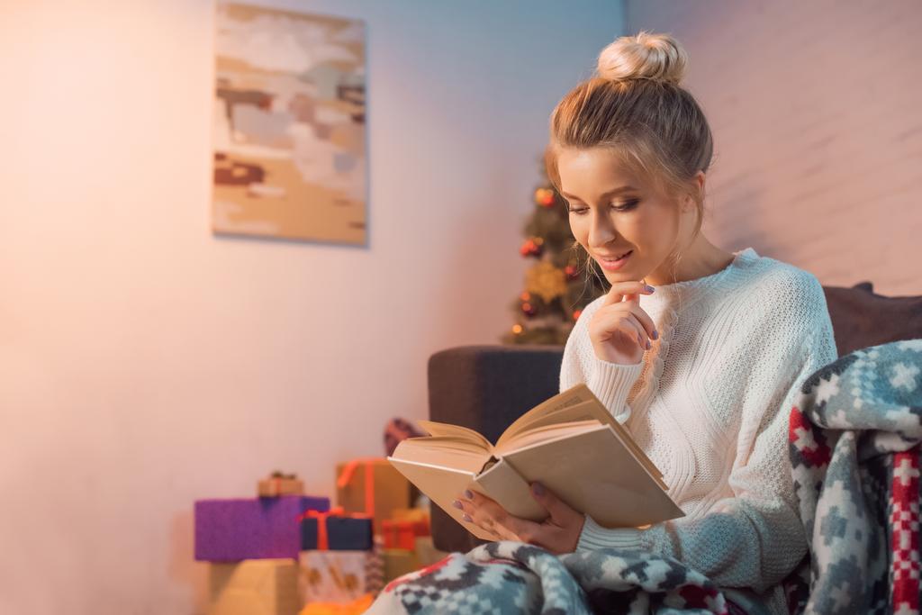 Chica encantadora sonriendo y leyendo libro en casa en la víspera de Navidad
 - Foto, imagen