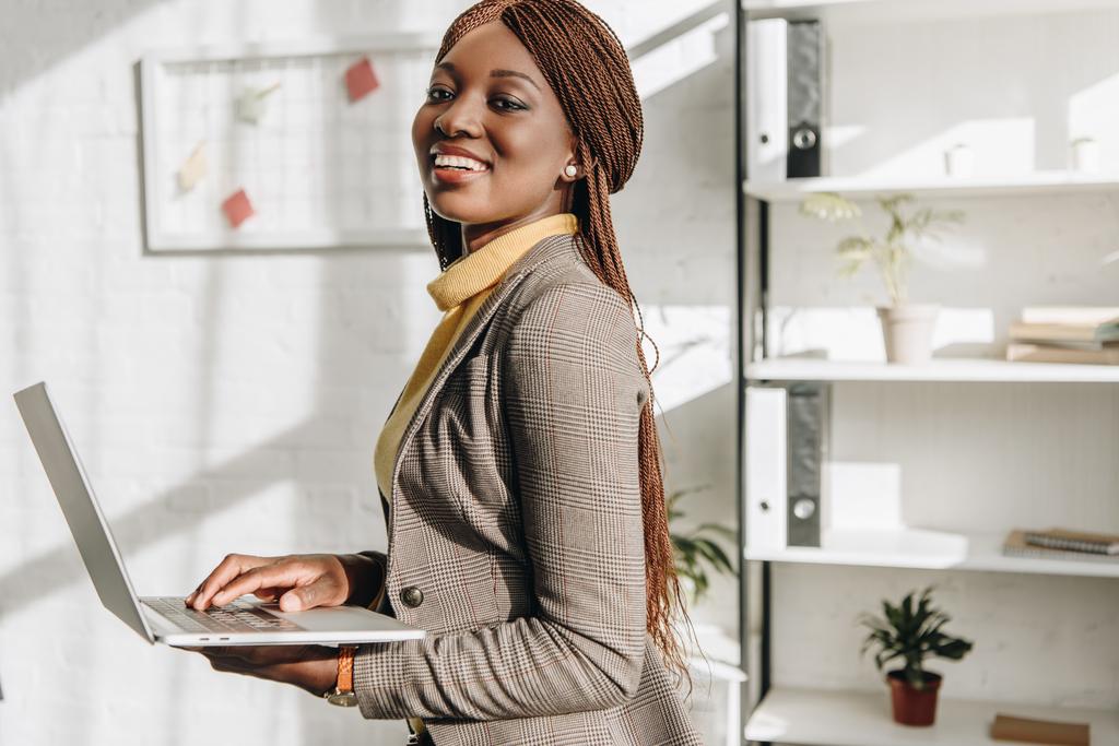 afro-amerikai felnőtt üzletasszony laptop tartja a munkahelyen, és mosolyogva néz a kamera - Fotó, kép