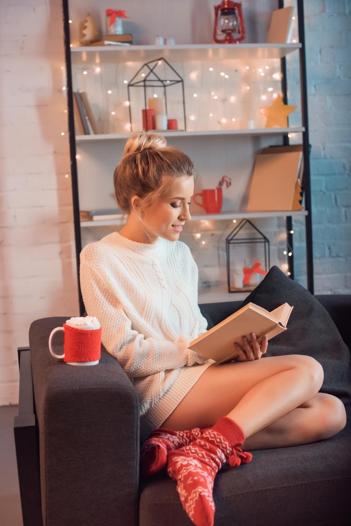 Mladá blondýnka s šálkem horké kakao a marshmallows sedí na gauči a čtení knih v době Vánoc - Fotografie, Obrázek