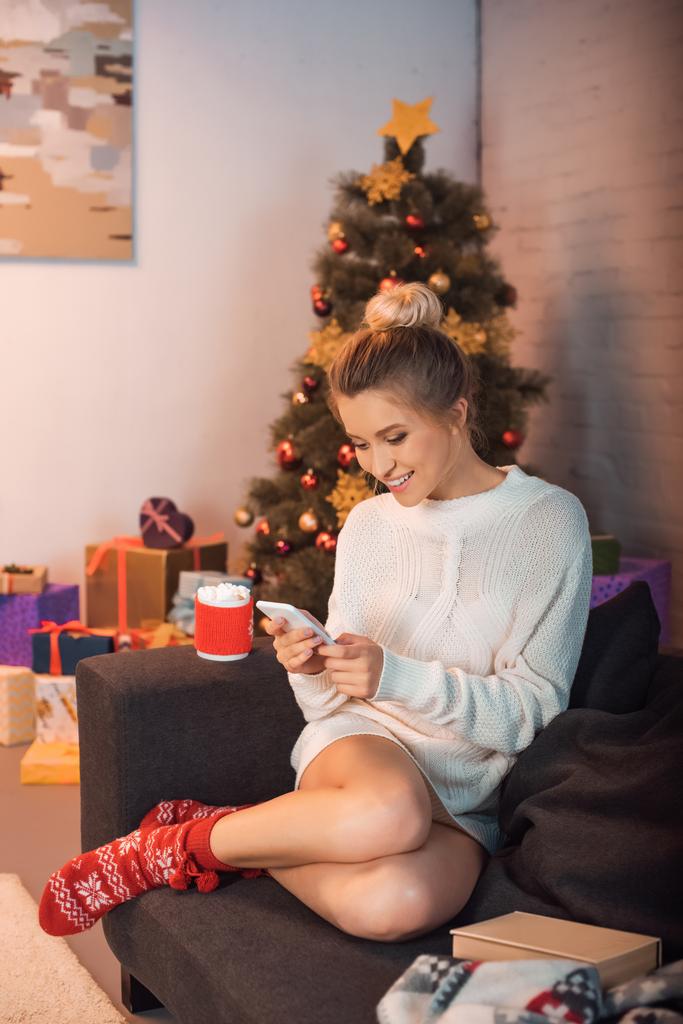 usměvavá mladá blond žena sedí na gauči a pomocí smartphone v době Vánoc - Fotografie, Obrázek