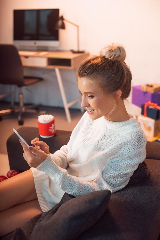 jeune femme blonde souriante assise sur le canapé et utilisant un smartphone à Noël
  - Photo, image