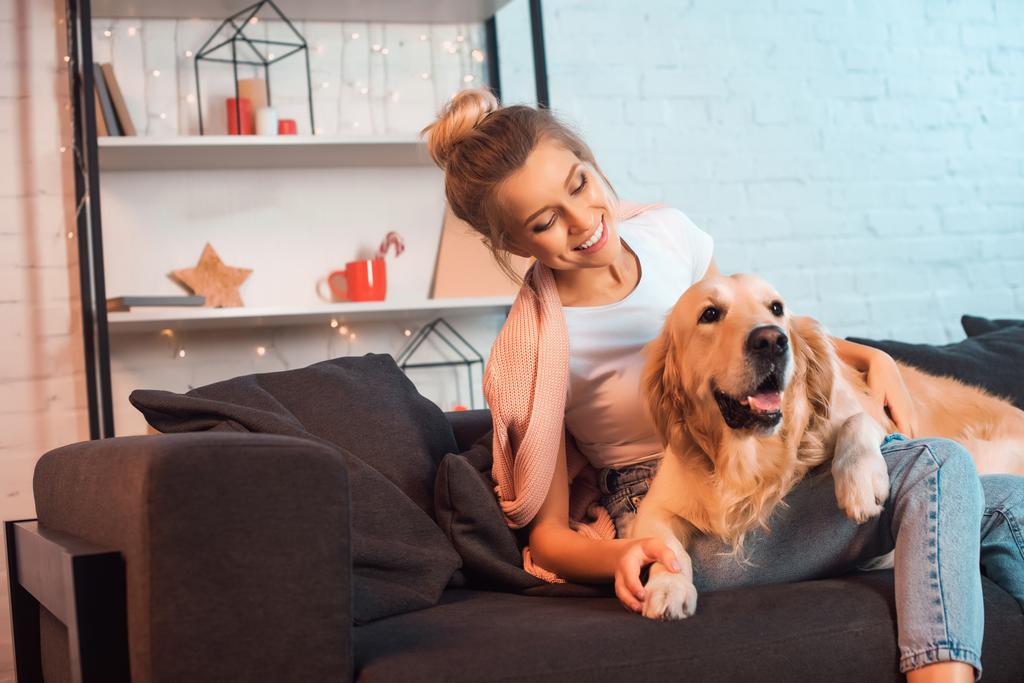 beautiful smiling young blonde woman sitting on couch and hugging golden retriever dog at christmas time - Photo, Image