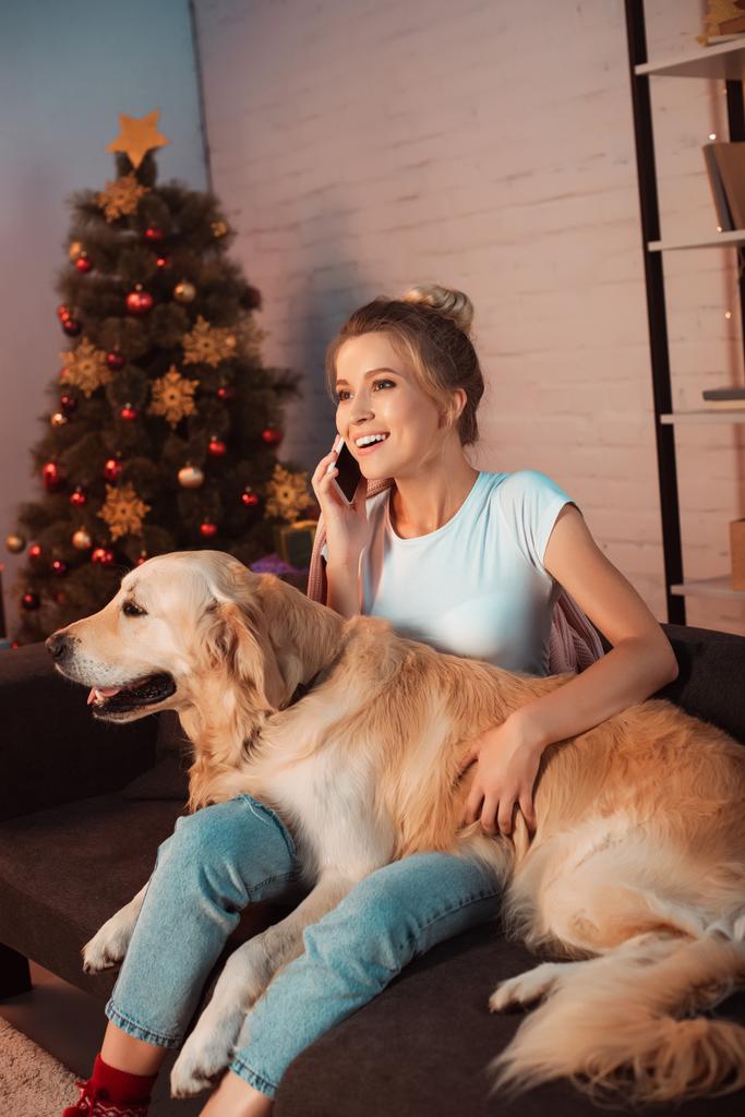 beautiful smiling young blonde woman sitting on couch with golden retriever dog and talking on smartphone at christmas time - Photo, Image