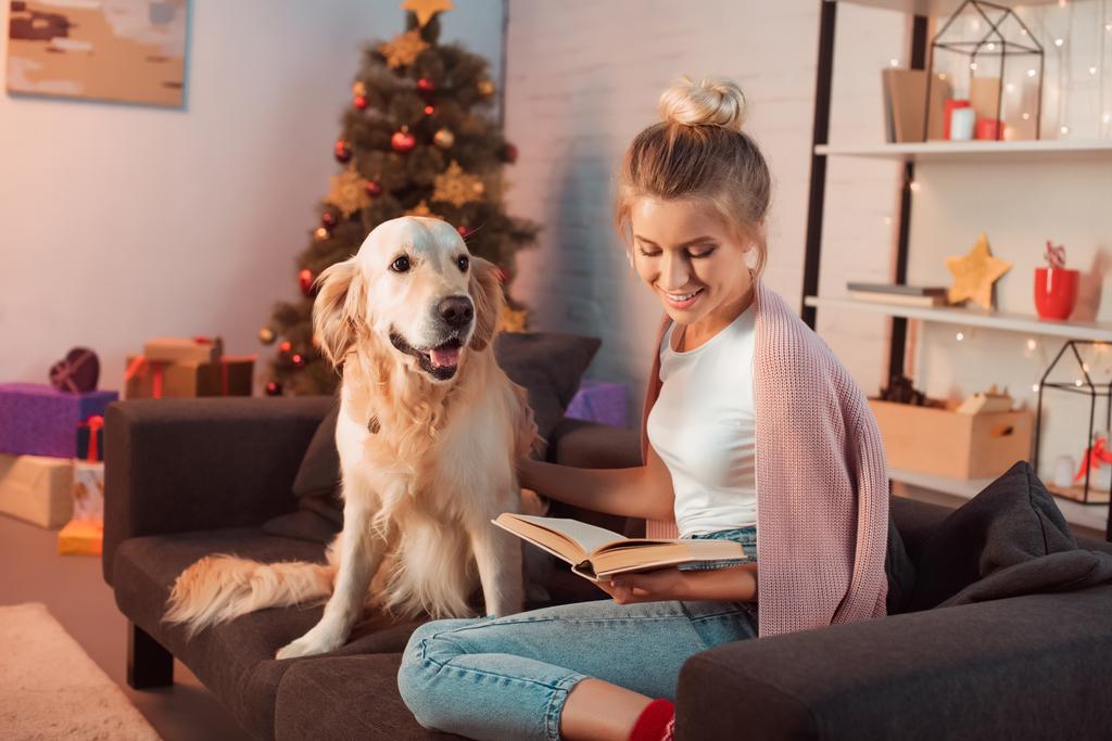 beautiful young blonde woman sitting on couch with golden retriever dog and reading book at christmas time - Photo, Image