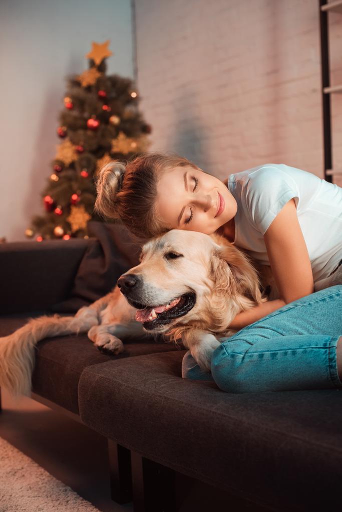 hermosa sonriente joven rubia en el sofá abrazando perro golden retriever en Navidad
 - Foto, Imagen