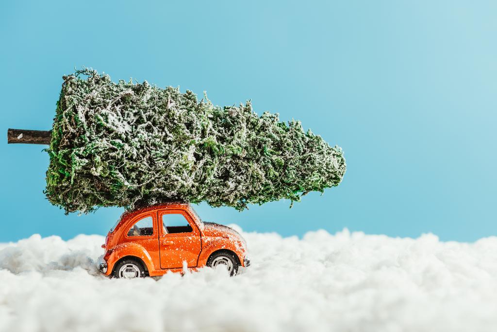 vista lateral del vehículo de juguete con árbol de navidad en miniatura en la azotea montado por la nieve de algodón sobre fondo azul
 - Foto, Imagen