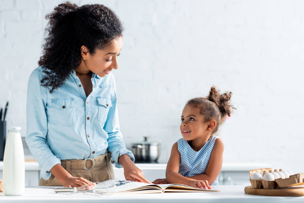 allegra madre e figlia afro-americana che si guardano, libro di cucina sul tavolo in cucina
 - Foto, immagini