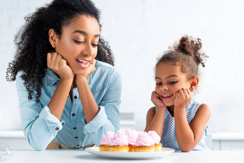 cheerful african american mother and daughter resting chins on hands and looking at cupcakes on table in kitchen - Photo, Image