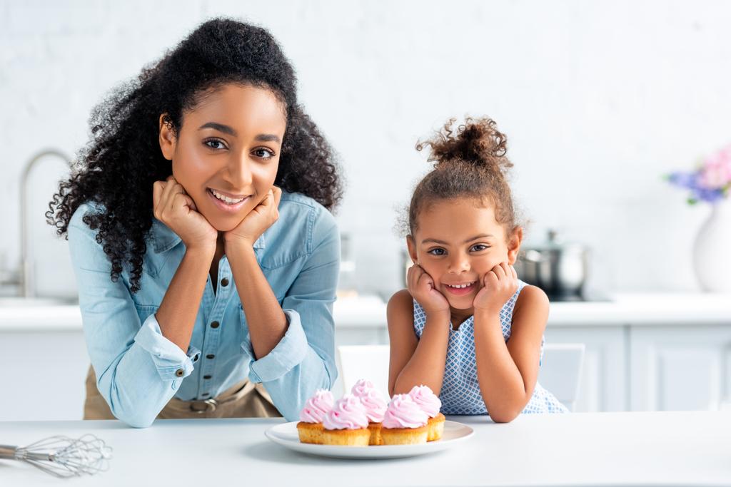vidám afro-amerikai anya és lánya álluk nyugszik a kezét, és látszó-on fényképezőgép, cupcakes táblázat konyha - Fotó, kép