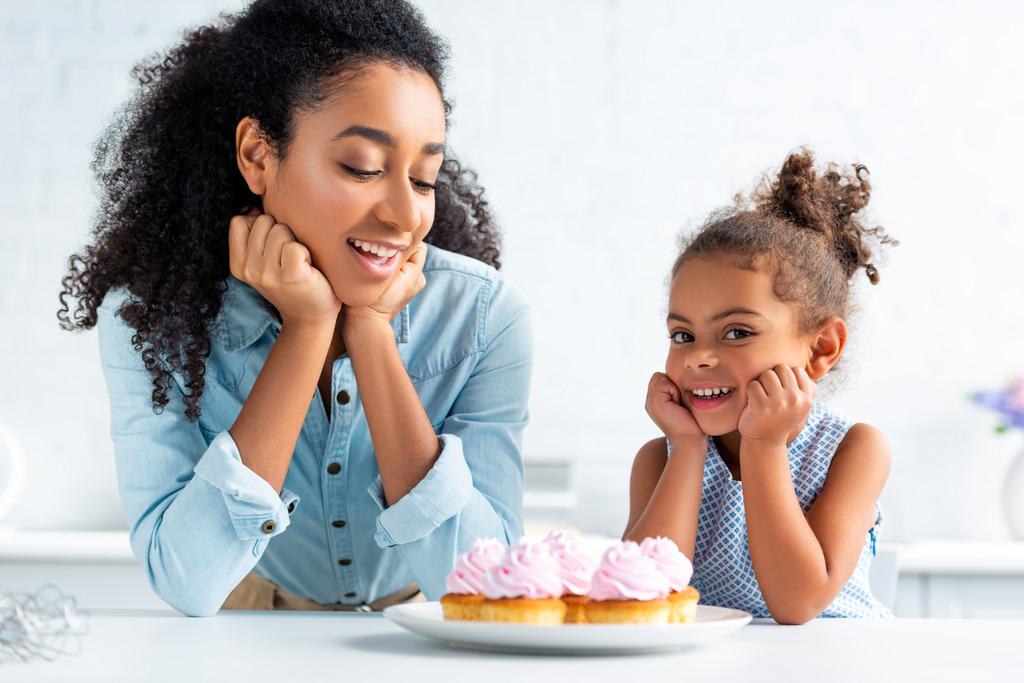 アフリカ系アメリカ人の母と娘のキッチンのテーブルでカップケーキ近く手にあごを安静時 - 写真・画像