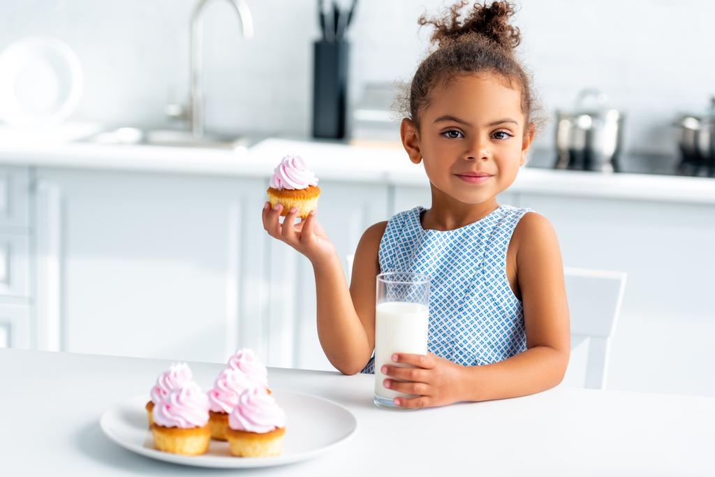 schattig Afro-Amerikaanse kind holding cupcake en glas melk in keuken - Foto, afbeelding