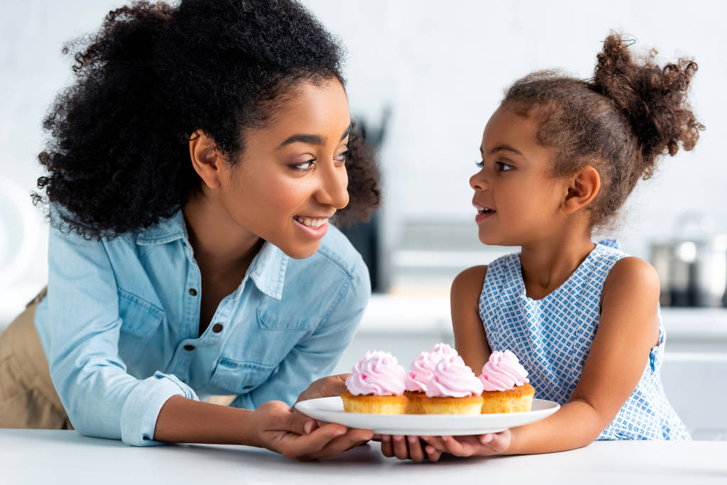 glimlachend Afro-Amerikaanse moeder en dochter zelfgemaakte cupcakes op te houden en kijken elkaar in de keuken - Foto, afbeelding