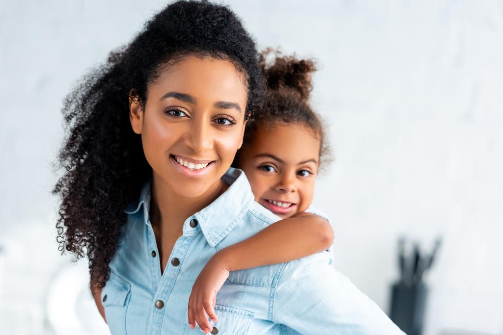 cheerful african american mother giving piggyback to adorable daughter in kitchen - Photo, Image