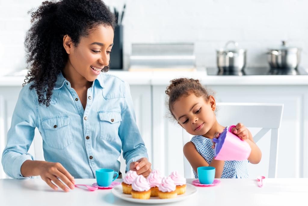 african american daughter and mother playing with plastic dishes in kitchen - Photo, Image