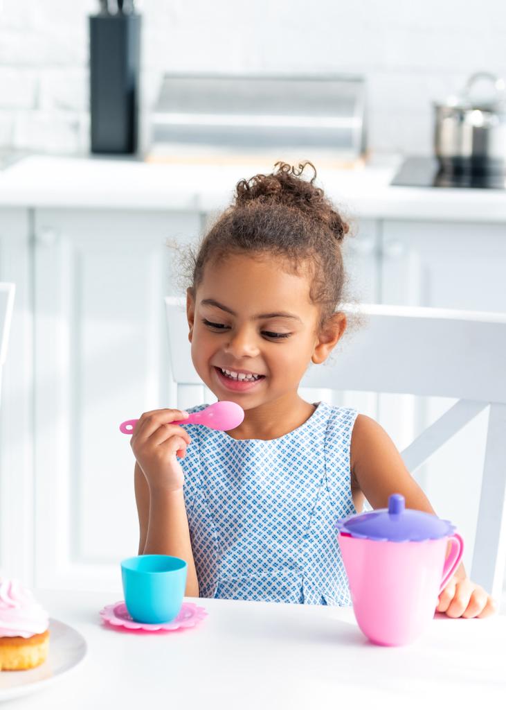 キッチンにプラスチック製の食器と座っているアフリカ系アメリカ人の子供の笑顔が愛らしい - 写真・画像
