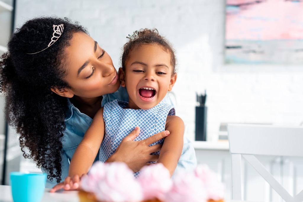 afro-americana mãe abraçando animado filha na mesa com cupcakes na cozinha
 - Foto, Imagem