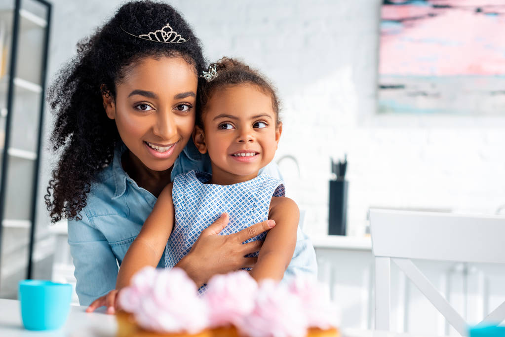 ελκυστική αφρικανική αμερικανική μητέρα αγκαλιάζει κόρη στο τραπέζι με cupcakes στην κουζίνα - Φωτογραφία, εικόνα