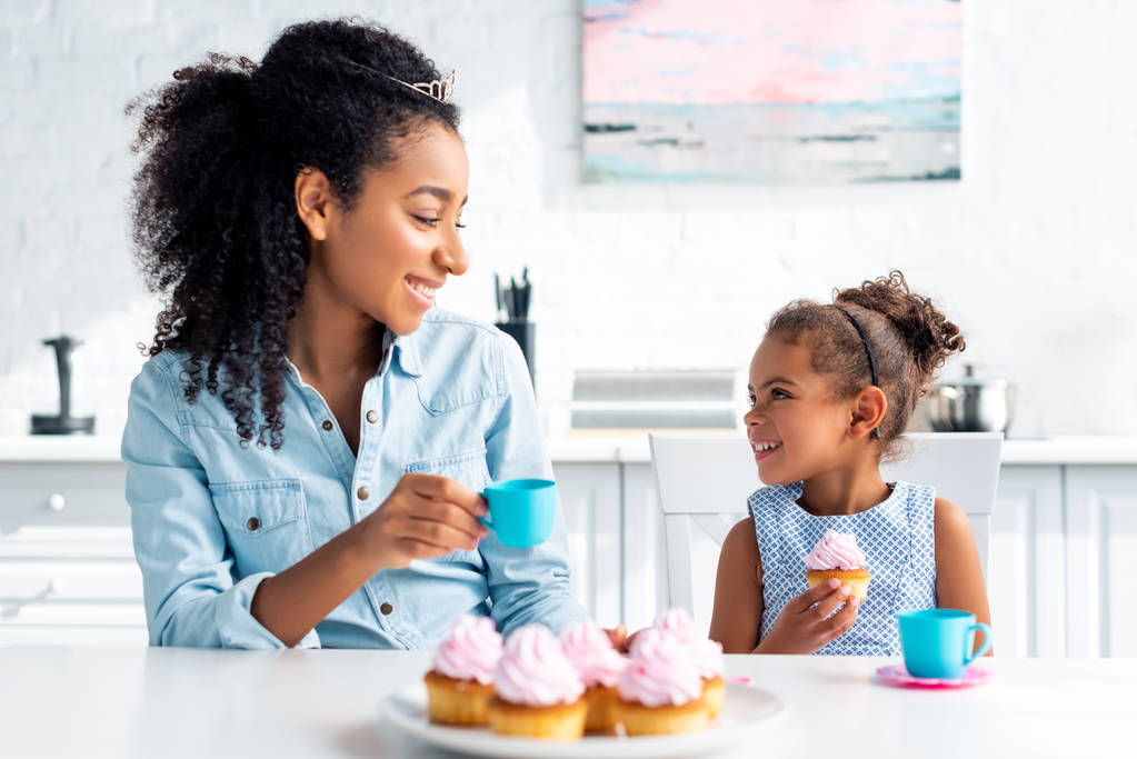 lachende Afro-Amerikaanse moeder en dochter met tiaras holding plastic beker en cupcake in keuken - Foto, afbeelding