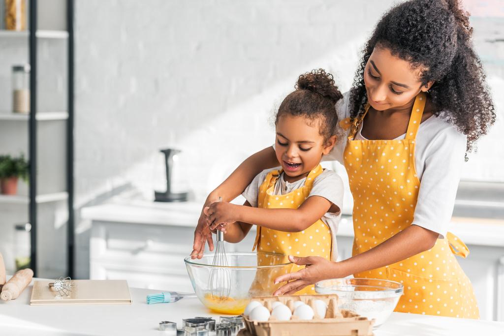Αφρικανική αμερικανική μητέρα και κόρη με κίτρινο χρώμα τα αυγά χτυπώντας για ζύμη στην κουζίνα ποδιές - Φωτογραφία, εικόνα