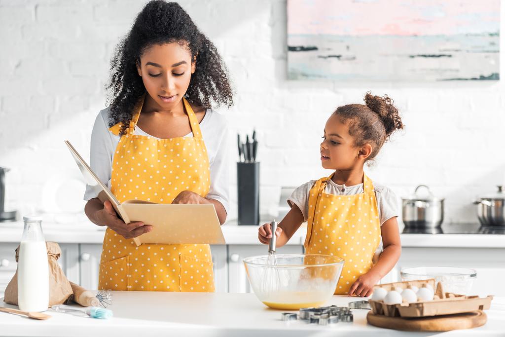 Африканская американская мать читает поваренную книгу и дочь готовит тесто на кухне
 - Фото, изображение